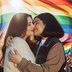 Two significantly overweight women, one of Caucasian descent with long brown hair and the other of South Asian descent with a short black bob hairstyle, embracing in a passionate kiss against a backdrop of a colorful LGBT flag fluttering in the wind. The atmosphere is celebratory and peaceful, with gentle sunlight beaming down upon them and the flag. Their expressions radiate love and acceptance, their clothes are casual yet stylish. There's an aura of positivity and harmony encapsulating the scene.